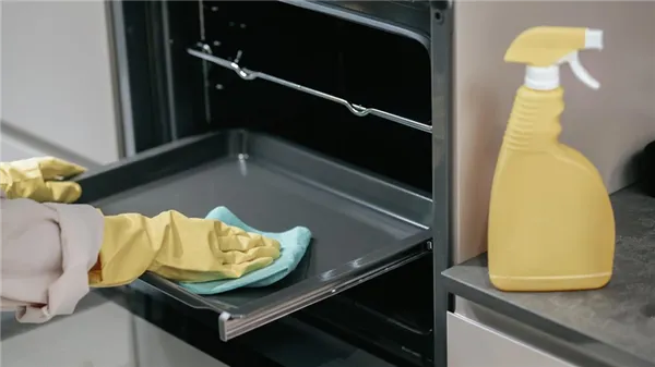 Открытую духовку вытираюь салфеткой. НА руках одеты желтые перчатки, рядом с духовкой стоит желтый пульверизатор